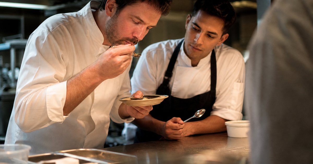 A chef tasting a new dish from a small plate at the pass of a restaurant kitchen while a sous chef watches closely holding a tasting spoon, heat lamps glowing above