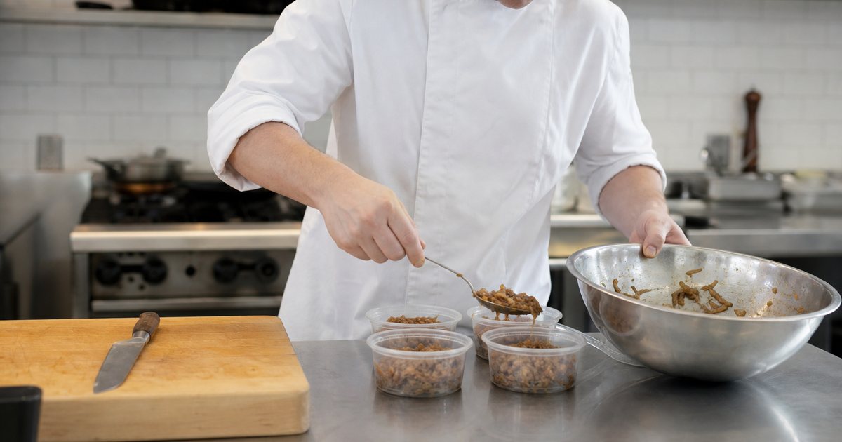 A chef in a white coat portioning caramelized onions from a large stainless mixing bowl into several smaller clear containers on a stainless steel prep table, knife and wooden cutting board beside, bright kitchen light from above