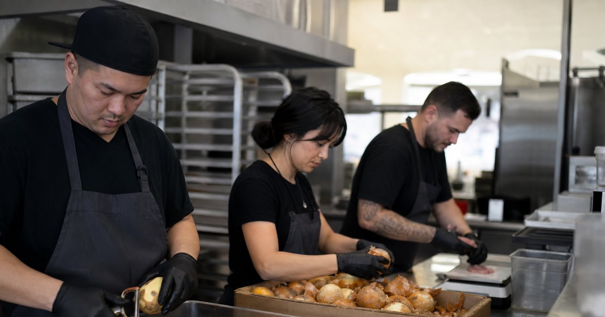 Three cooks working side by side at a long stainless prep table in a restaurant kitchen, one peeling potatoes into a bin, one breaking down a case of onions, one portioning proteins on a scale